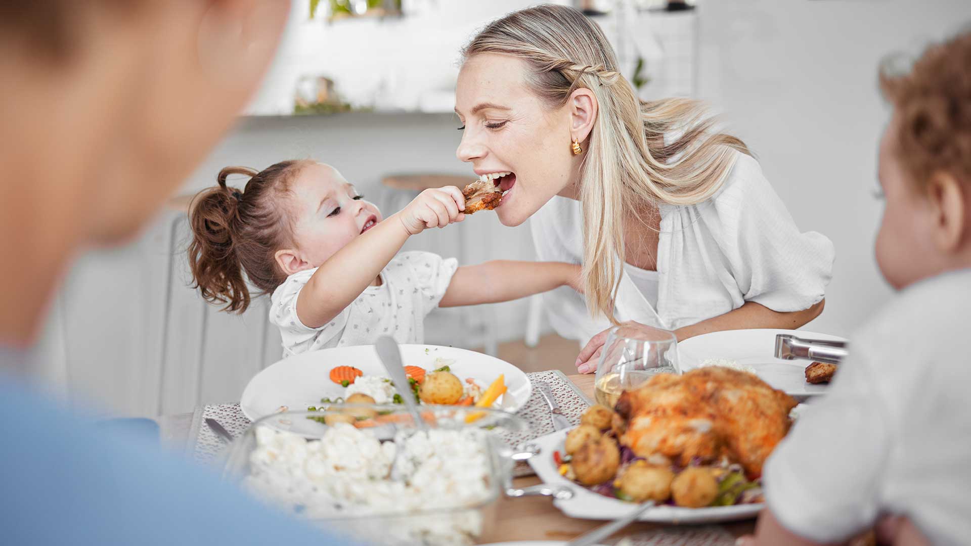 Eine kleine Tochter reicht ihrer mutter eine Hähnchenkeule. Die Mutter beisst herzhaft hinein.