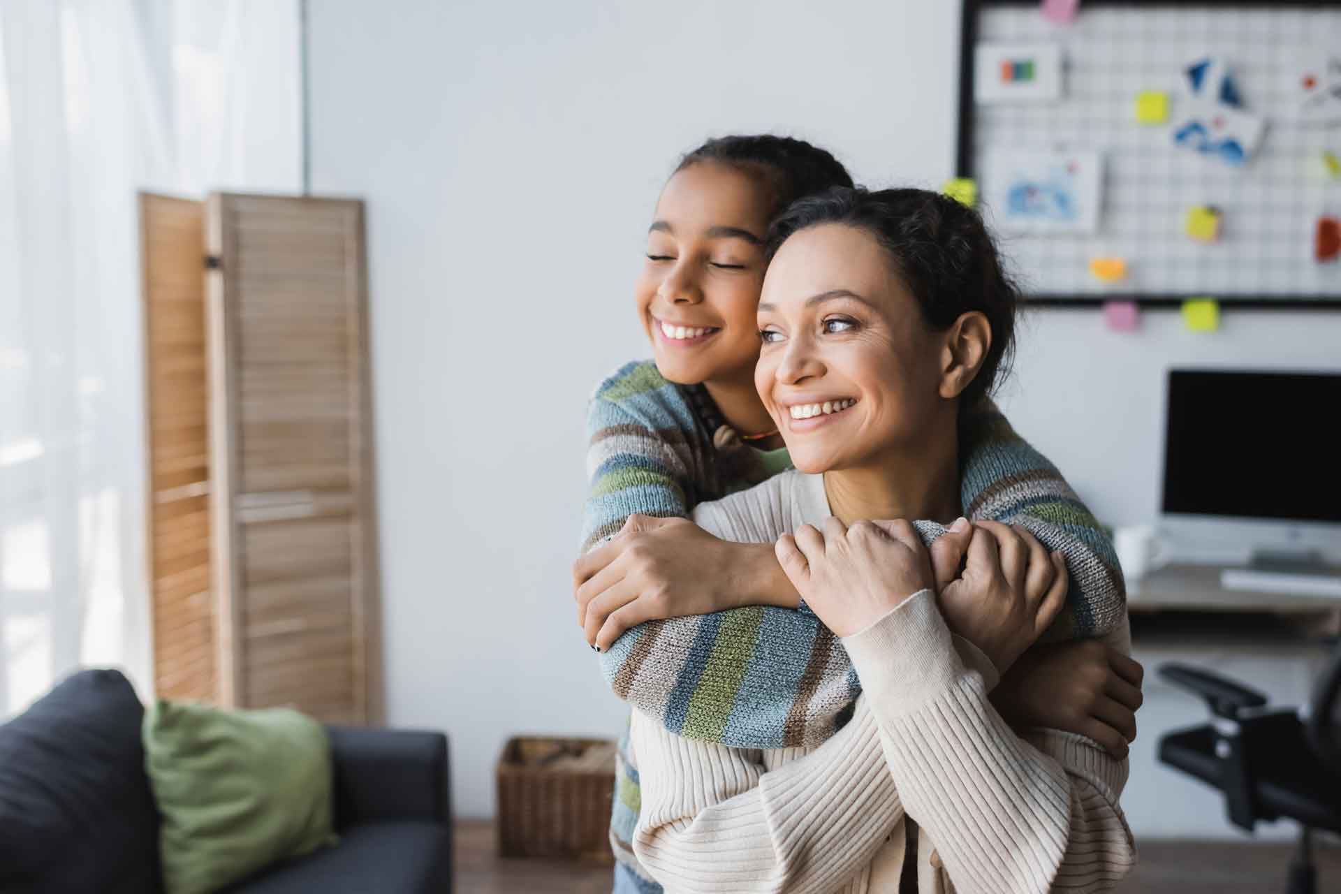 Mutter und Tochter lächeln und blicken aus dem Fenster. Die Tochter umarmt die Mutter von hinten.