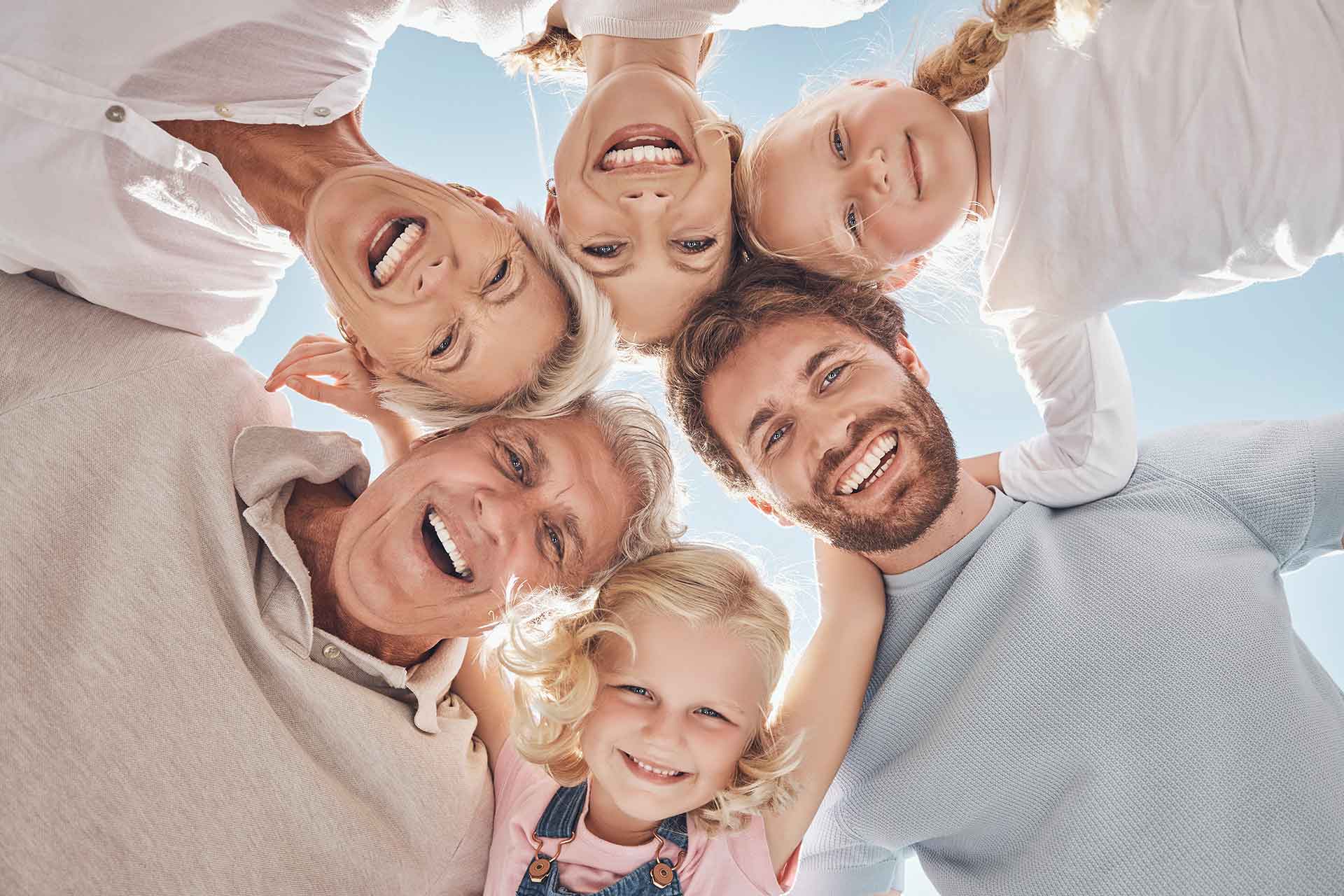 zahnarztpraxis-hlavacek-hero-prophylaxe Eine 6-köpfige Familie blickt nach unten in die Kamera und alle strahlen mit offenen Mündern in die Kamera. Hinter ihnen ist strahlend blauer Himmel zu sehen.