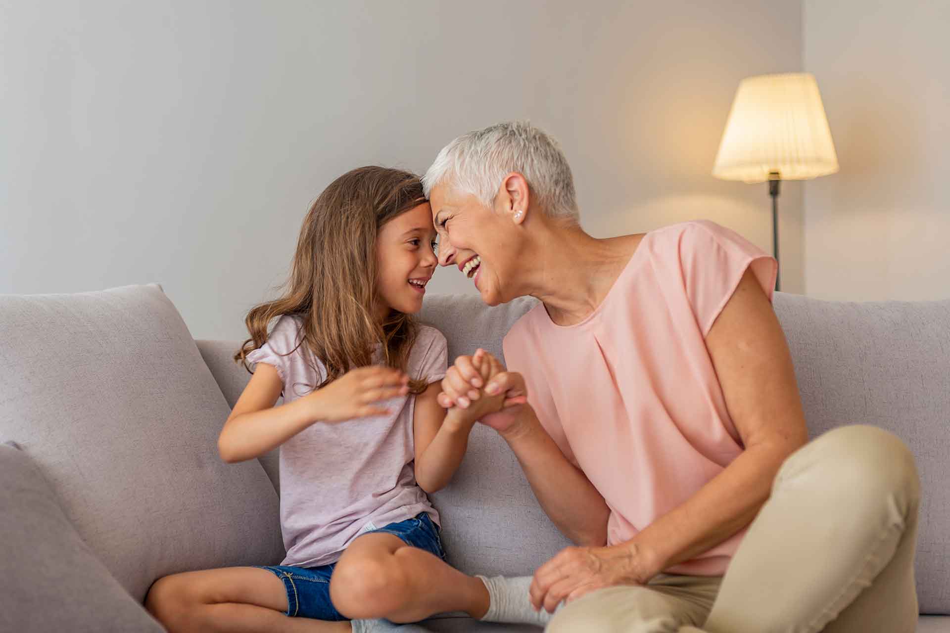 Eine Großmutter und ihre Enkelin sitzen auf dem Sofa und lachen sich gegenseitig an.