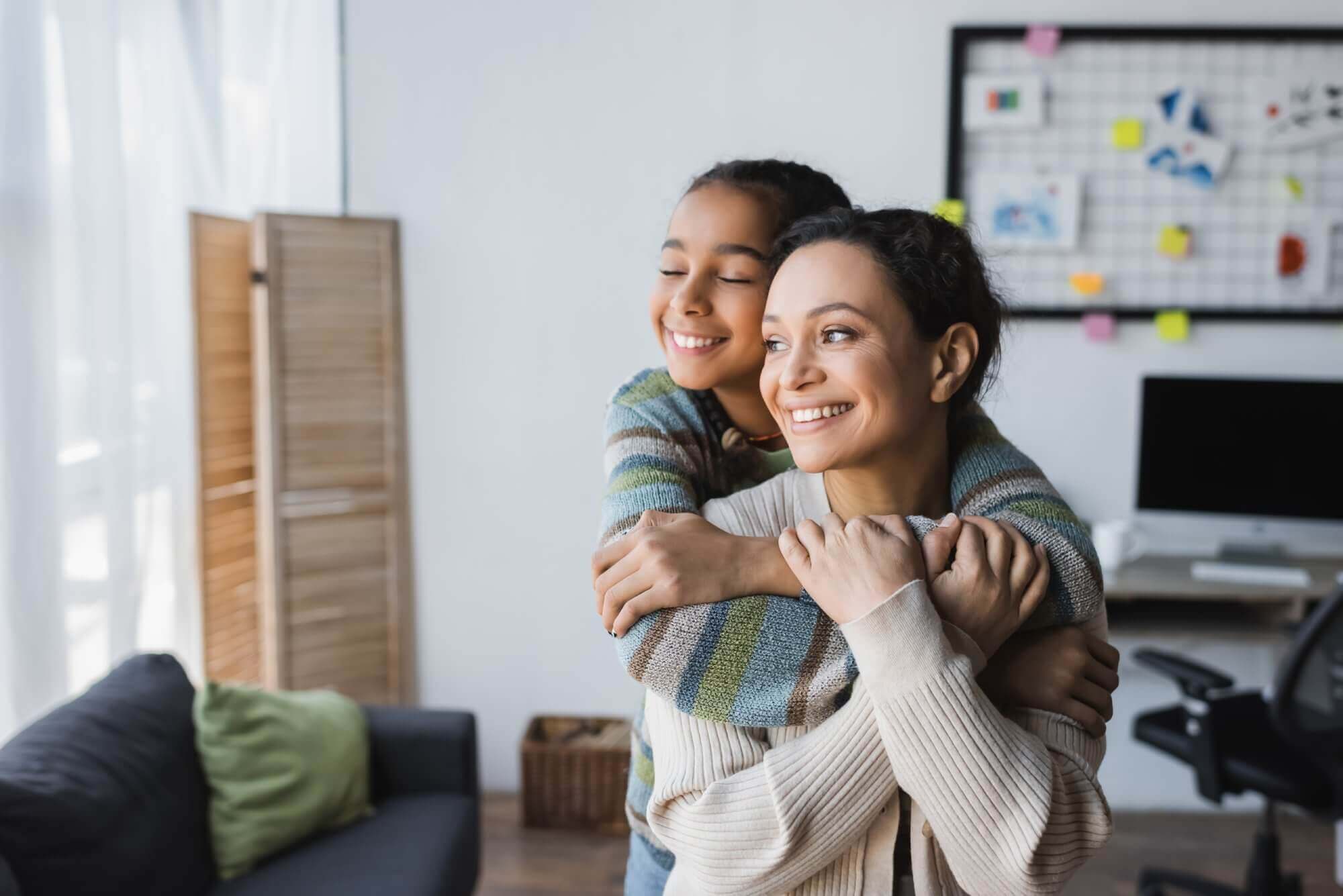 Mutter und Tochter lächeln und blicken aus dem Fenster. Die Tochter umarmt die Mutter von hinten.